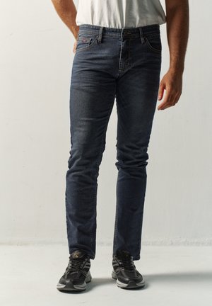 Hombre con vaqueros ajustados azul oscuro, camisa blanca metida por dentro y zapatillas negras, de pie frente a un fondo claro y sencillo.