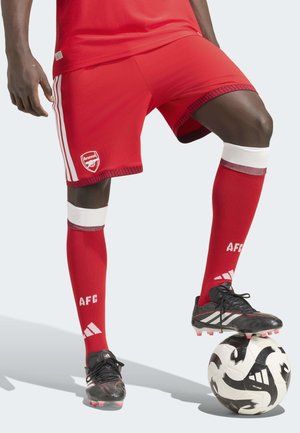 Piłkarz w czerwonych szortach i skarpetach Arsenalu balansuje jedną stopą czarno-białą piłkę nożną Adidas.