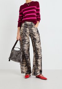 Personne portant un pull bordeaux avec des rayures roses, un pantalon large à motif tigre, des ballerines rouges à bout pointu, tenant un sac à main gris foncé.