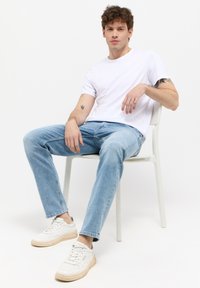 Tricou alb cu maneci scurte, blugi de culoare albastru deschis si adidasi albi cu talpa bej. Barbatus stă pe un scaun, prezentând o ținută casual.
