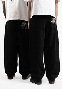 Baggy black denim pants with back pockets featuring white spider web embroidery. Fabric has a smooth texture, designed for a relaxed fit.