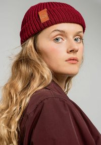 Burgundowa dziana czapka z teksturowanym wzorem i brązową skórzaną łatą. Długie, faliste włosy częściowo widoczne pod czapką.