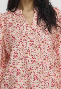 Tissu à motif floral en rose doux avec des accents rouges et jaunes. Présente un col à patte et des manches bouffantes pour un ajustement décontracté.