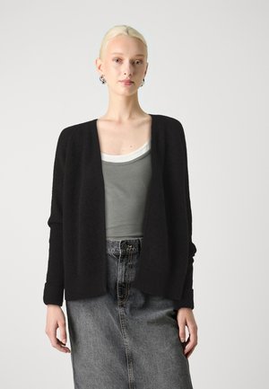 Črn, teksturiran kardigan z odprtim sprednjim delom in dolgimi rokavi, oblečen preko sivega tank topa, v kombinaciji z jeans krilom z visokim pasom.