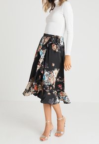 Schwarz floral gemusterter Rock mit Rüsche am Saum, der lebendige orangefarbene, blaue und weiße Blumen zeigt, kombiniert mit einem weißen Oberteil und silbernen Riemchenschuhen.