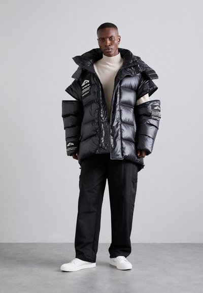 Doudoune noire avec un col montant, coupe oversize et finition brillante. Présente des logos et textes en évidence sur les manches. Associée à un pantalon noir et des chaussures blanches.