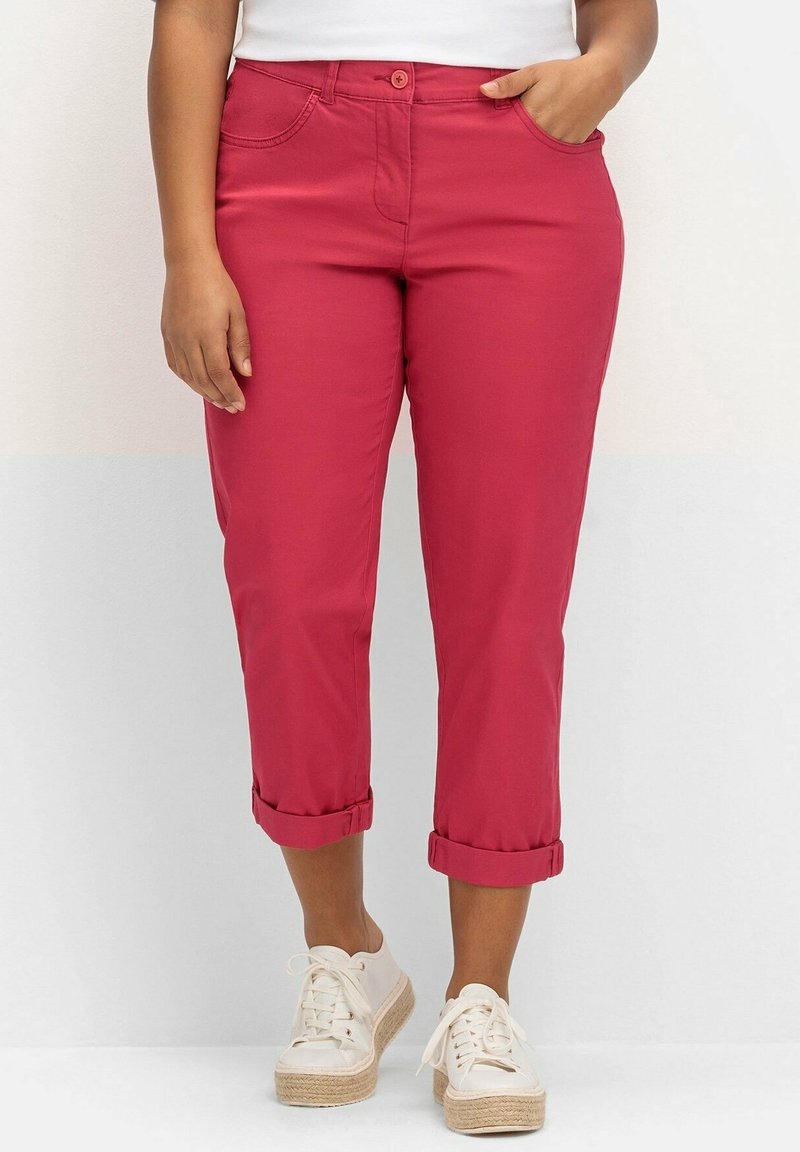 Personne portant un pantalon court framboise retroussé, des baskets blanches et un haut blanc, se tenant devant un fond blanc uni.
