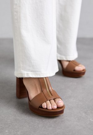 Brūni ādas platformas papēži ar rāvējslēdzēja aizdari. Noapaļota pirksta forma, ko papildina koka līdzīga laukuma papēdis. Balti platajā kājās daļēji redzami.