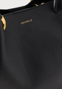 Τσάντα χειρός από μαύρο δερματίνι με υφή, με χρυσούς χειρολαβές σε σχήμα δακτυλίων και χρυσό λογότυπο "COCCINELLE" στο πάνω μπροστινό μέρος.