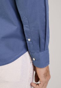 Lichtblauwe shirt van katoenstof, met een knoopmanchet met twee witte knopen en vervaardigde plooi-details langs de mouw.