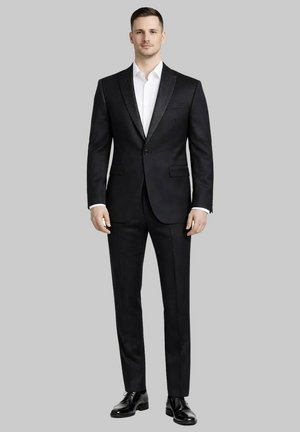 Homme debout en smoking noir à col châle, chemise blanche, chaussures habillées noires, sur fond gris uni.