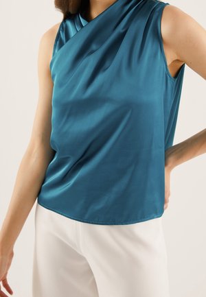 Vrouw draagt mouwloze teal satijnen top en witte broek met één hand op de heup tegen een effen achtergrond.