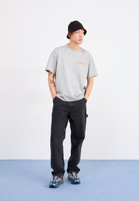 Gray short-sleeve t-shirt with orange "DICKIES" logo, paired with baggy black pants and black knit bucket hat, worn with multicolor sneakers.