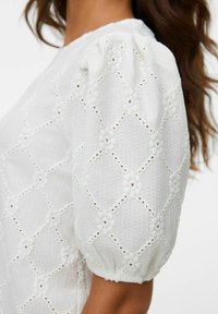 Hvit bluse med strukturert stoff med diamantmønstre og blomsterdetaljer. Korte pufferm med elastiske mansjetter.