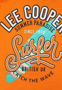 T-shirt en coton orange avec un texte audacieux en noir et blanc, un graphique de planche de surf verte avec des accents rayés, et des phrases décoratives.