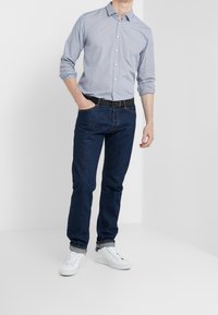 Hombre con camisa azul clara de botones y mangas arremangadas, jeans azul oscuro con puños arremangados, cinturón negro y zapatillas blancas de pie contra un fondo blanco.