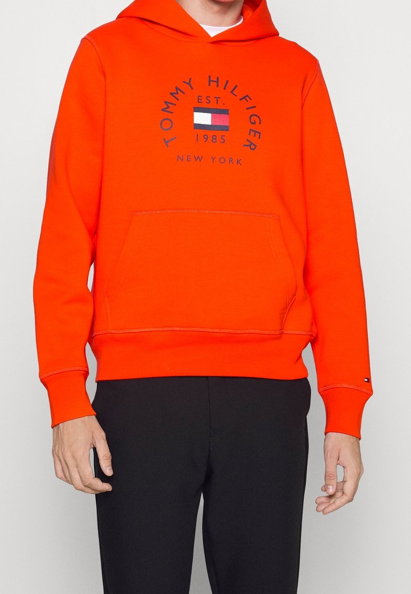 Oranje hoodie met een voorvak, voorzien van een cirkelvormig logo met de tekst "Tommy Hilfiger" en een vlagdesign, gemaakt van een gladde, gestructureerde stof.