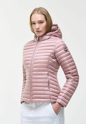 Frau trägt eine hellrosa gesteppte Kapuzenjacke mit Frontreißverschluss und Händen in den Taschen einer weißen Hose, vor einem schlichten hellen Hintergrund.