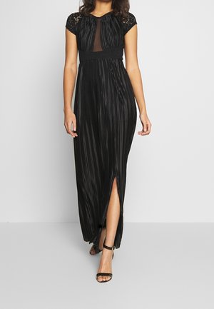 Mujer vestida con vestido maxi negro plisado con mangas de encaje y abertura frontal, combinado con sandalias negras de tacón alto con tiras.