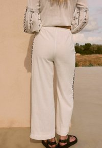 Pantaloni larghi di lino bianchi con accenti laterali ricamati neri. Orlo piatto e cintura elasticizzata. Indossati con sandali marroni scuri.