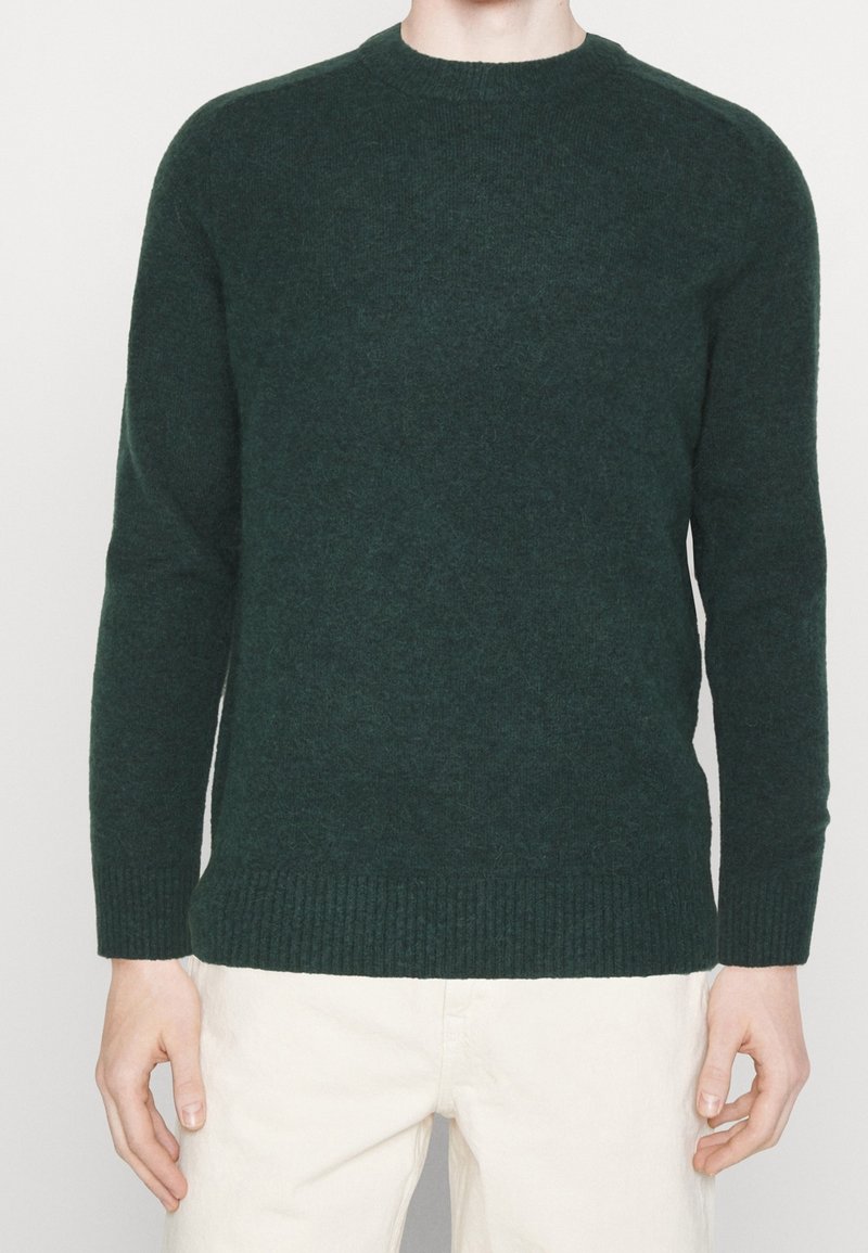 Man in een donkergroene gebreide trui met lange mouwen en geribbelde manchetten en zoom, gecombineerd met lichtgekleurde broek, staande tegen een witte achtergrond.