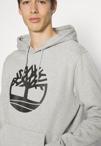 Grijze hoodie van zacht materiaal met een groot zwart logo van een boom en een gebogen lijn onderaan. Verstelbare trekkoordcapuchon.