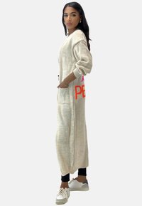 Długi beżowy sweter kardigan z grubym splotem, z oversize'owymi rękawami, bocznymi kieszeniami i wyrazistym różowo-pomarańczowym napisem "PEACE" po jednej stronie.