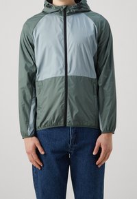 Veste légère à capuche en vert foncé et gris clair, avec poignets élastiques, fermeture éclair à l'avant et texture lisse et brillante.