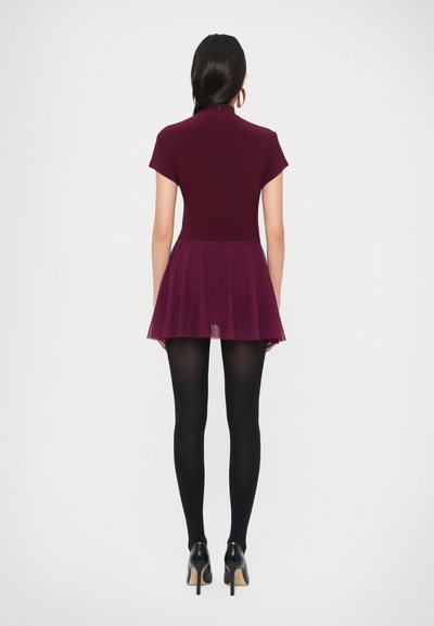 Βουργουνδί φόρεμα σκάτερ με εφαρμοστό μπούστο, κοντά μανίκια και φουντωτή διάφανη φούστα από μουσελίνα, συνδυασμένο με μαύρες καλτσοδέτες και ψηλά τακούνια.