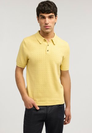 Jonge man met donker krullend haar draagt een geel korte mouw polohemd met kleine perforaties en donkere broek, staand met één hand in de zak.