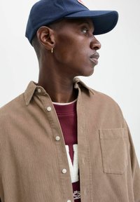 Brązowa koszula z korduroy z zapięciem na guziki i kieszenią, nałożona na bordowy sweatshirt z białym napisem, noszona z granatową czapką.