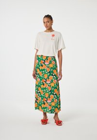 Witte top met korte mouwen en rood logo, gecombineerd met een lange groene rok met roze rozen en oranje patronen. Rood strik-sandalen met hak.