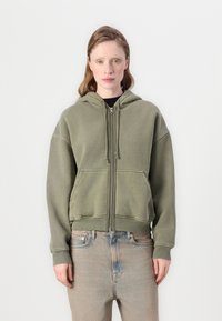 Maslinasto zelena zip-up dukserica s kapuljačom na vezanje, prednjim dzepovima i rebrastim manžetama. Mekana tkanina s opuštenim dizajnom.