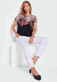 Zwarte top met kleurrijke bladachtige patronen, witte broek en zwarte flats. Soepele stof, korte mouwen en een relaxte pasvorm. Eenvoudige armband als accessoire.