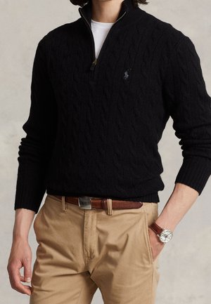 Mand iført en sort kabelstrikket sweater med kvart-lynlås over en hvid skjorte, beige bukser med brunt bælte og et armbåndsur på venstre håndled.