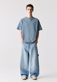 Blå, kortærmet t-shirt parret med oversized lyseblå denimjeans. Jeansene har en løs pasform og for lomme. Grå sneakers med tan-såler.