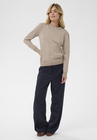 Pull en maille beige avec un col rond et des poignets côtelés, associé à un pantalon bleu marine ample avec poches latérales et jambes larges.