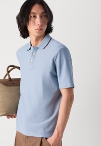 Hellblaues Polohemd mit strukturiertem Stoff, kurzen Ärmeln und einem kontrastierenden dunklen Kragen, gehalten mit einer braunen Handtasche.