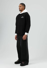 Sweat à capuche noir avec capuche doublée de blanc, arborant un logo sur la poitrine. Associé à un jean noir ample et des chaussures noires avec des accents roses.