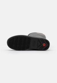 Botte en caoutchouc noire avec une semelle texturée présentant un accent circulaire rouge et un design côtelé diagonal. Parte supérieure lisse avec un bout arrondi.