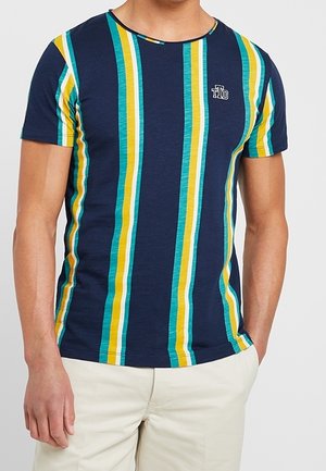 Mężczyzna w granatowym t-shircie z pionowymi żółtymi, turkusowymi i białymi paskami, zestawiony z jasnobeżowymi spodniami, dłonie swobodnie przy ciele.
