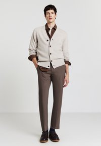 Beige gestrickter Cardigan mit dunklen Knöpfen, getragen über einem braunen Hemd mit Kragen. Braune maßgeschneiderte Hose und dunkle Schnürschuhe runden das Outfit ab.