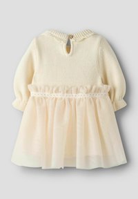 Strikket kremfarget livstykke med rund hals og knappelukking, kombinert med et lagdelt tyllskjørt i myk ferskenfarge. Detalj med rysjekant ved midjen.