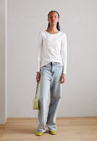 White long-sleeve shirt, light blue loose jeans, green bag, and matching green shoes. Plain background with wooden floor.