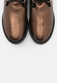 Botines metálicos de bronce con un acabado suave, que cuentan con detalles en negro y hardware dorado, punta redonda y suela de goma.