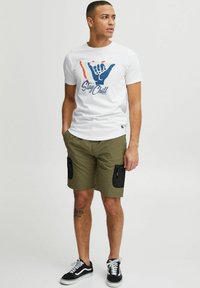 Hvid t-shirt med grafisk design, der viser en håndsign og teksten "Stay Chill." Olivengrønne shorts med dobbelte lynlåslommer. Sorte sneakers.