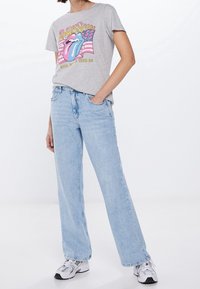 Persona vestida con una camiseta gráfica de los Rolling Stones en gris, pantalones vaqueros de pierna ancha en azul claro y zapatillas blancas, de pie con una mano en el bolsillo.