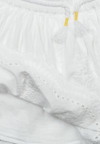 Prenda de algodón blanca con cintura elástica, adornada con bordados de flores y pequeños detalles de ojal, rematada con borlas amarillas.