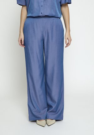 Personne portant un pantalon large bleu ample associé à des chaussures à bout pointu beige clair, debout devant un fond uni.