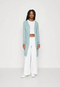 Lyseblå lang cardigan laget av mykt materiale, med åpen front, to sidelommer, og parret med en hvit crop top og vide bukser.
